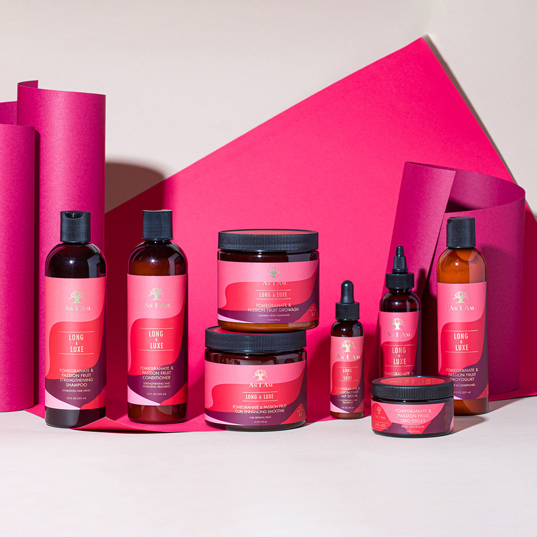 A collection of products from the As I Am Long & Luxe range, including shampoo, conditioner, leave-in conditioner, oils and jars, arranged in front of a pink background.