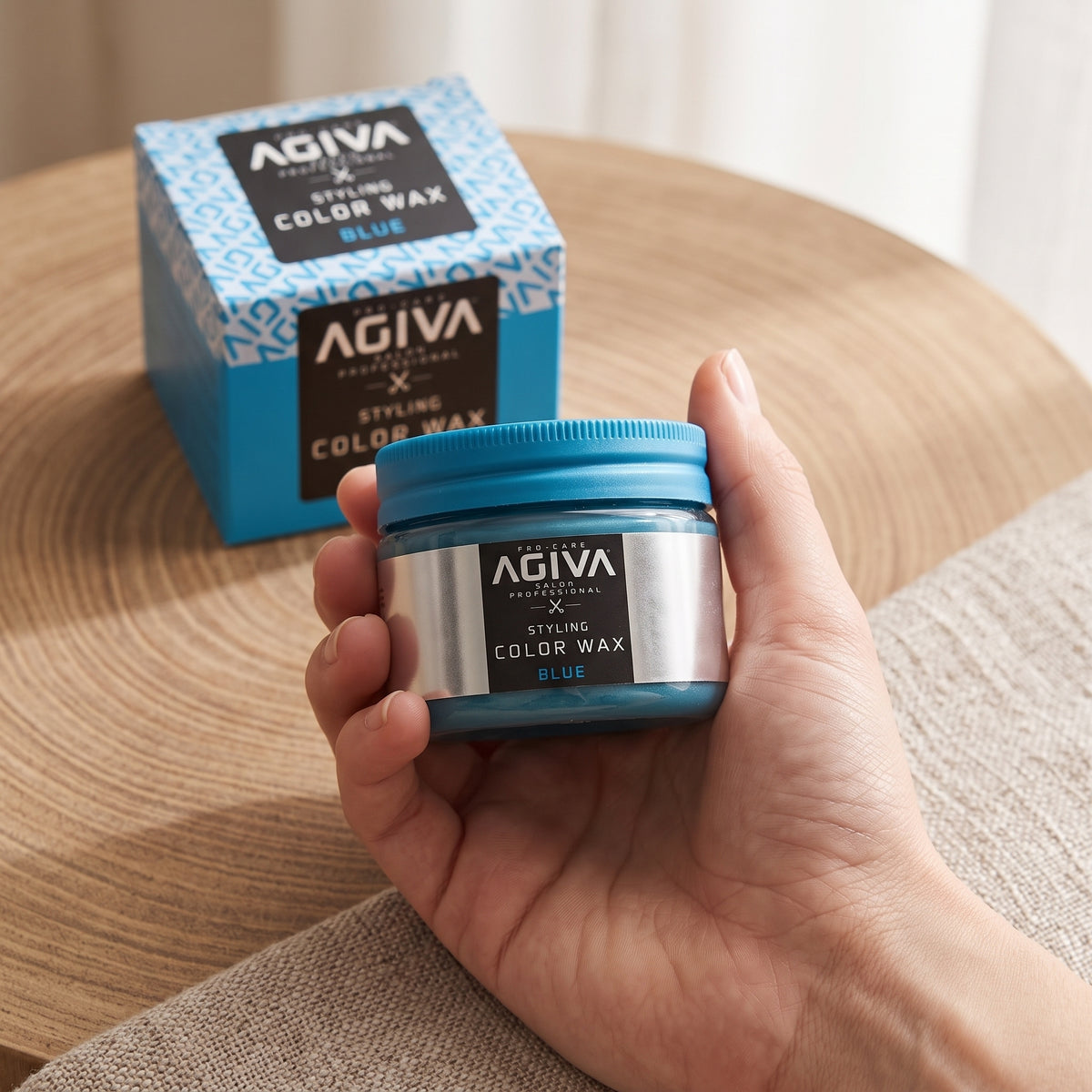 Hand holding AGIVA Styling Color Wax in blue color, with product box in background on wooden table.