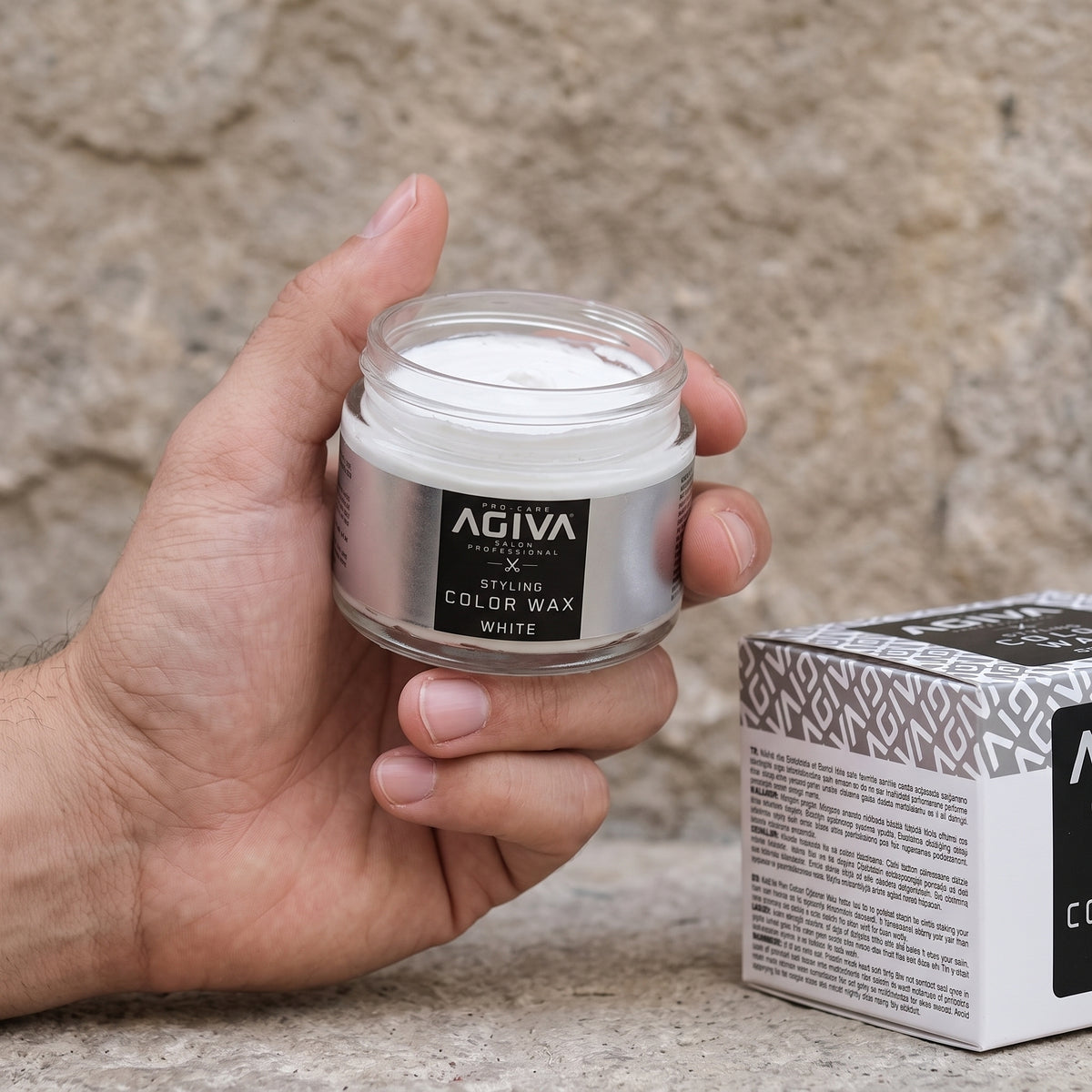 Hand holding open glass jar of Agiva Styling Color Wax White next to box on stone background.