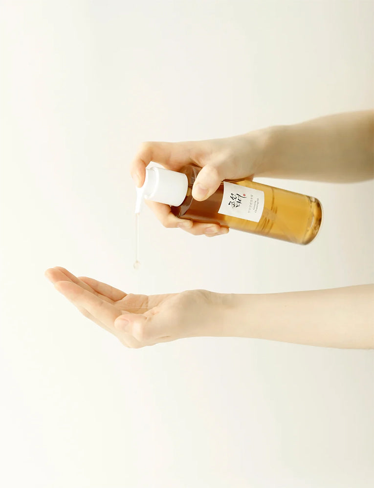 Beauty of Joseon Ginseng Essence Water 150ml is pumped into the palm of the hand from an amber bottle against a light, neutral background.