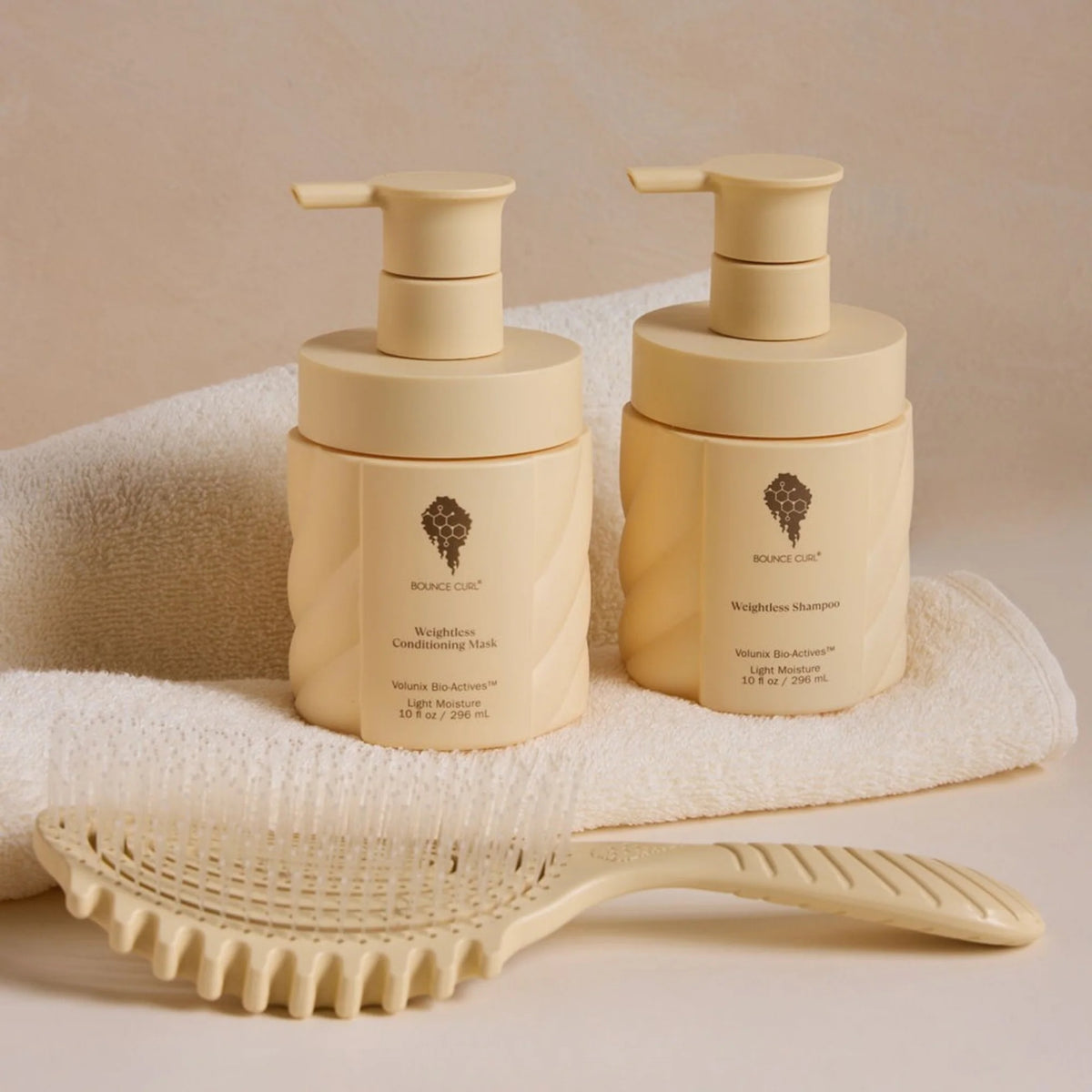 Bounce Curl Weightless Shampoo and Conditioning Mask in cream bottles with pump, placed on white towel next to hairbrush.