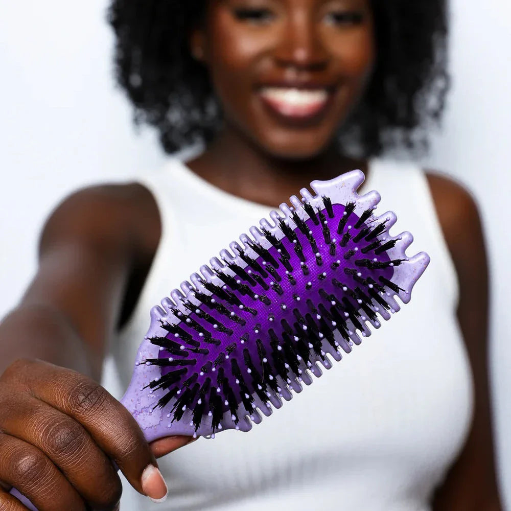 Woman with curly hair holds a purple Bounce Curl Volume EdgeLift Brush Purple close to the camera with a smile.