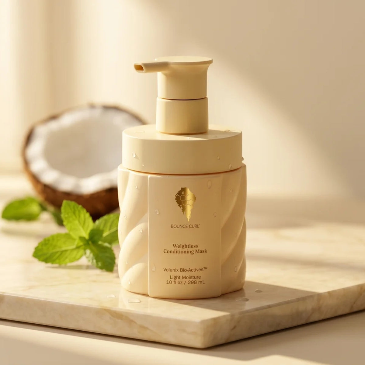 Bounce Curl Weightless Conditioning Mask in a cream colored pump bottle on a marble board, surrounded by coconut and fresh mint leaves.