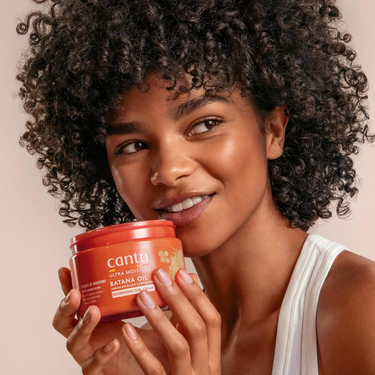 Woman with curly hair smiles and holds Cantu Ultra Moisture Batana Oil Nourishing Curl Cream to her cheek.
