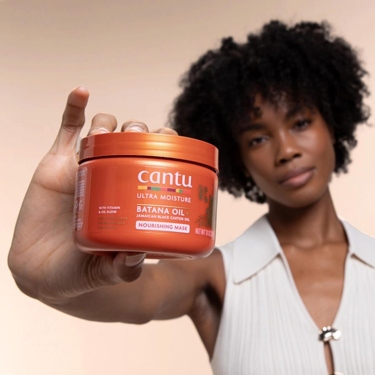 Woman with naturally curly hair holds Cantu Ultra Moisture Batana Oil Nourishing Hair Mask towards the camera.