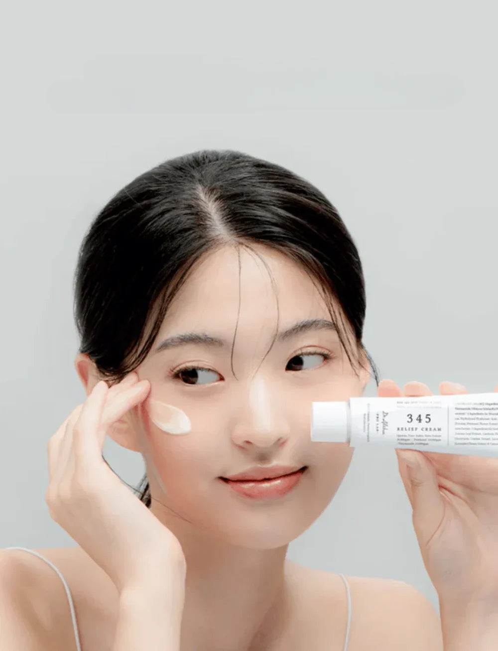 Woman with glowing skin applies Dr. Althea 345 Relief Cream 50ml to cheek with a gentle smile and holds tube in hand.