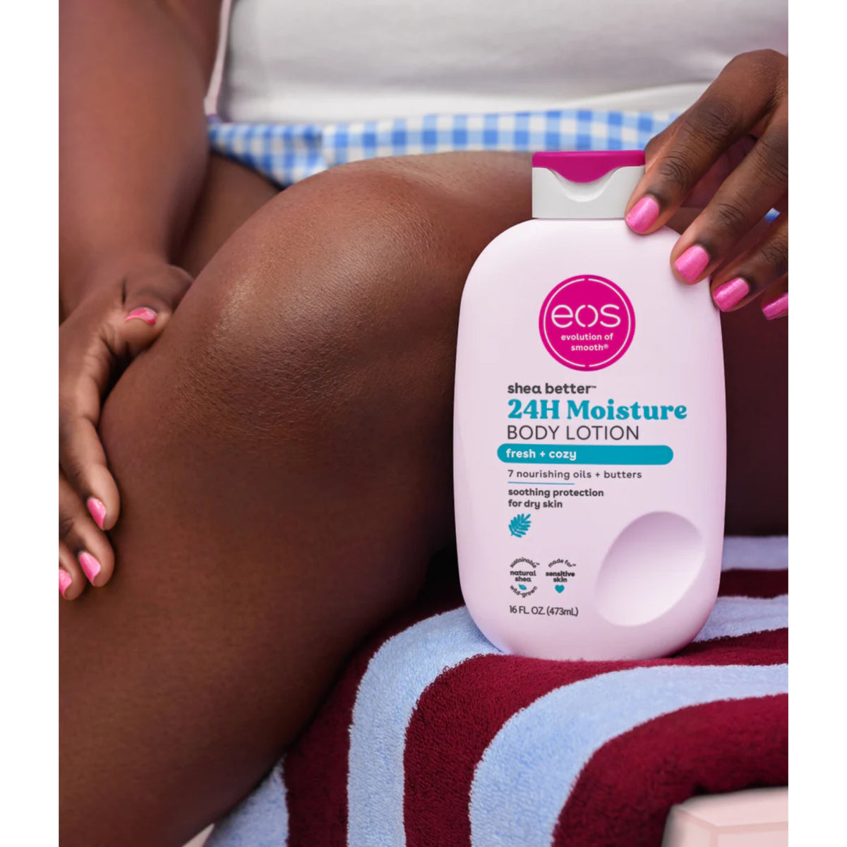 eos Shea Better 24H Moisture Body Lotion in white bottle with pink cap held up to tanned leg on striped towel.