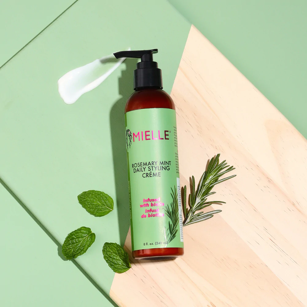 Mielle Rosemary Mint Multi-Vitamin Daily Styling Crème (240ml) is shown with a dollop of cream to demonstrate the consistency of the product. Highlights natural ingredients such as mint and rosemary.