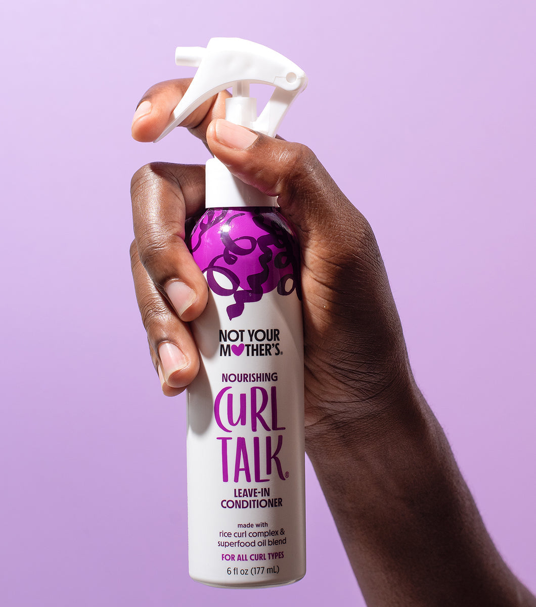 A dark hand sprays Not Your Mother's Curl Talk Nourishing Leave-In Conditioner in front of a solid purple background.
