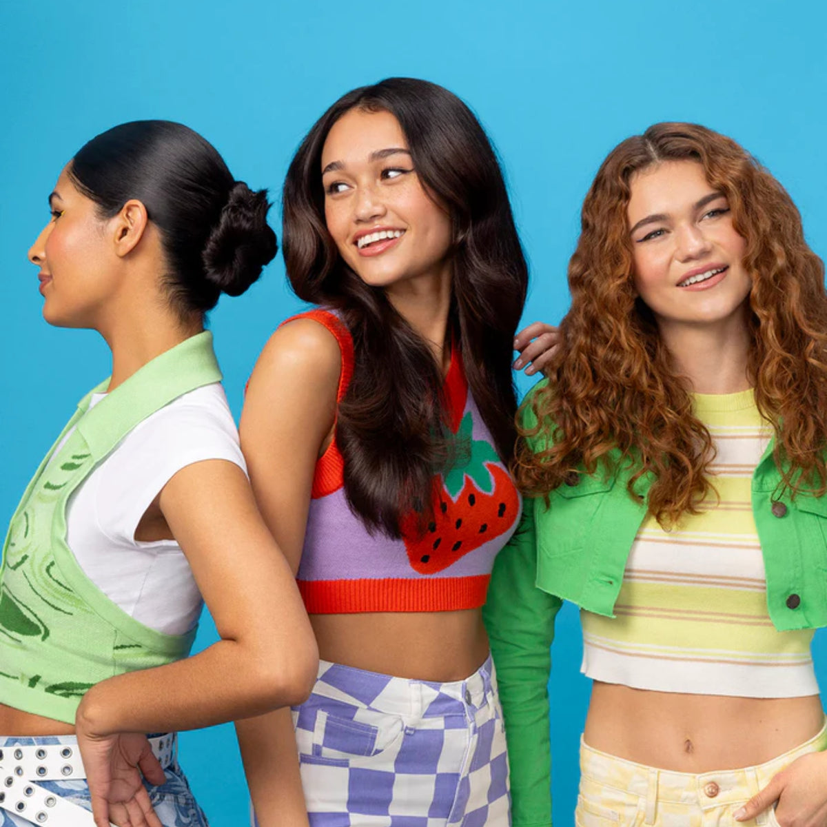 Three smiling young women in colorful summer clothes with jackets and crop tops against blue background.