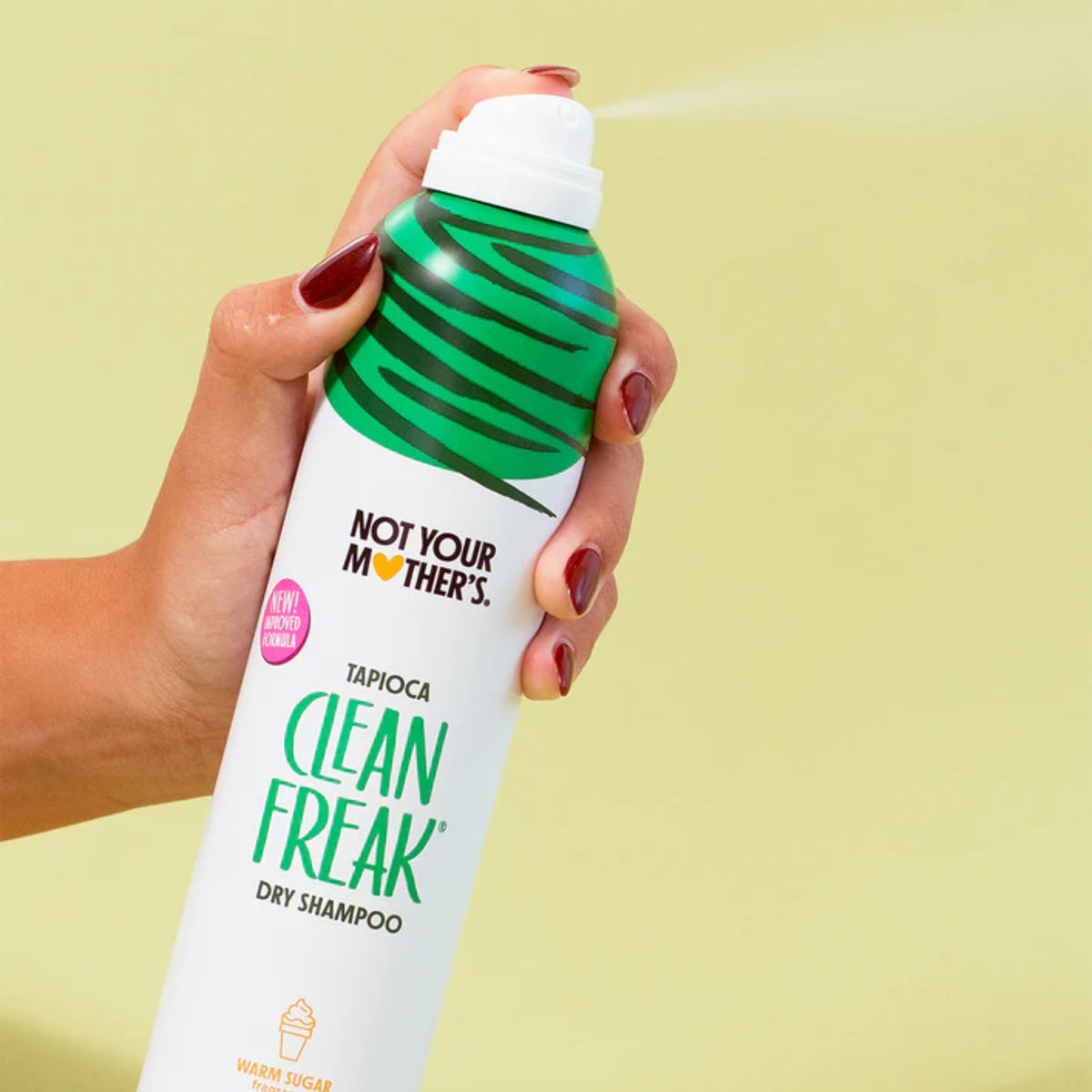 Hand holding Not Your Mother's Clean Freak dry shampoo in green can sprayed against green background.