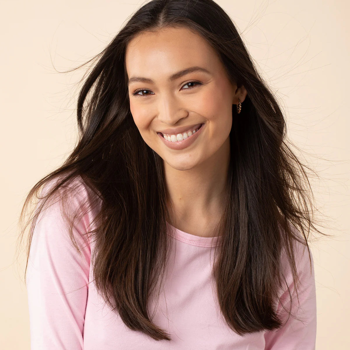 Smiling woman with long dark brown hair wearing pink long sleeve blouse and gold earrings against beige background.