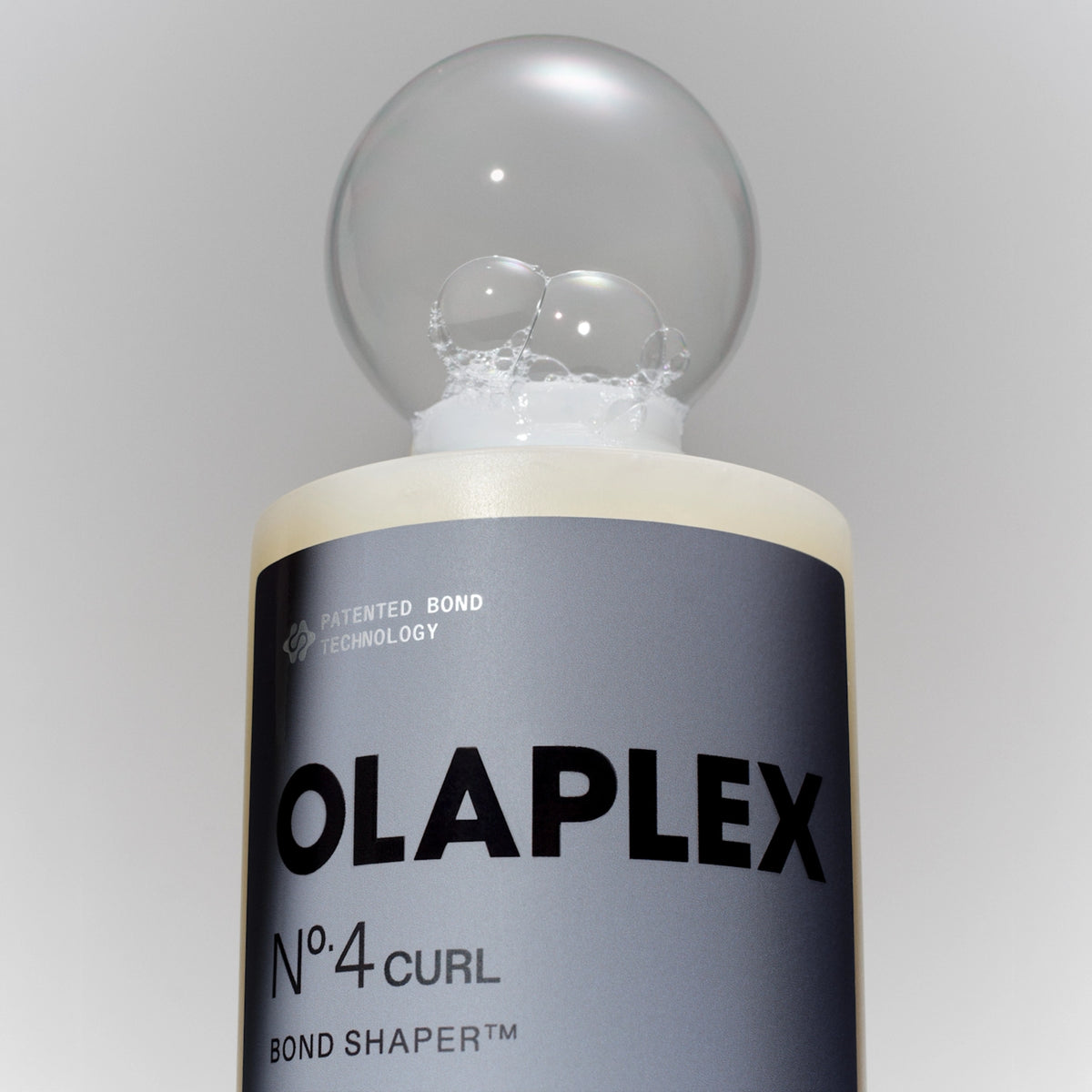 Olaplex No.4 Curl Bond Shaper with transparent ball cap and foam on the top of the bottle against a gray background.